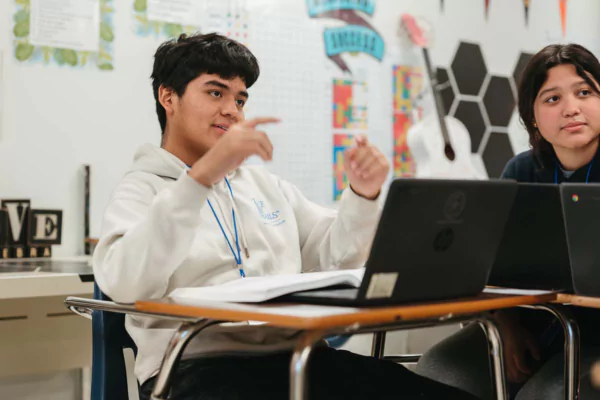 A high school aged student is using MATHia at a laptop and smiling as he talks with someone off camera.