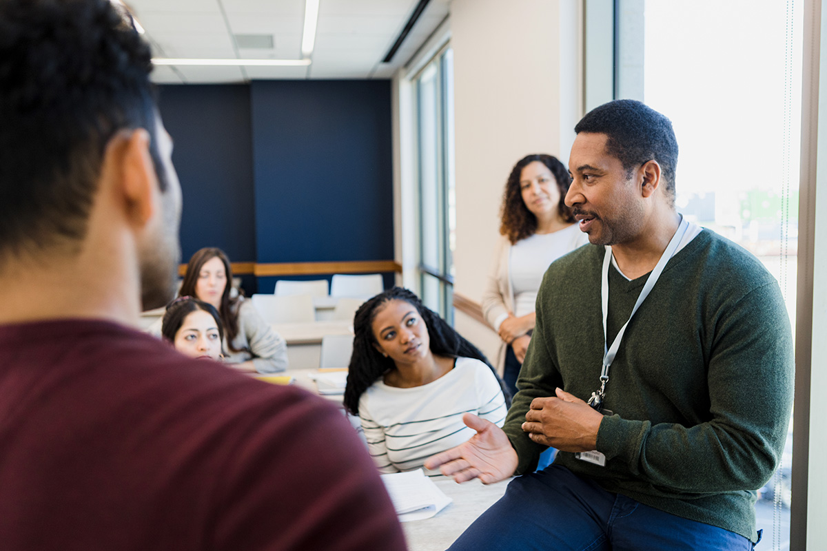 A teacher with a lanyard discusses the 2024 AP Chief Reader reports in a bright meeting room. Seated around the table, a female teacher in a white shirt and others listen attentively as natural light streams through the large windows, illuminating their focused expressions.