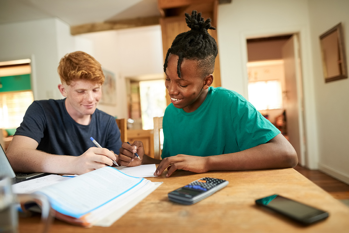 Two students sit at a table engaged in homework; the one on the right writes, while the other observes with a pen. Books, a calculator, and a smartphone are scattered around as they experience productive struggle in math, fully immersed in their focused home setting.