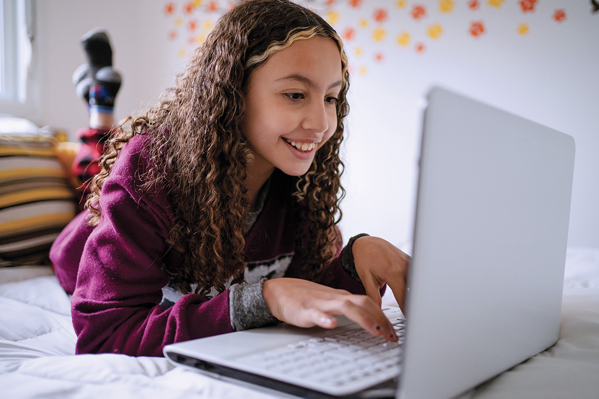 A young person with curly hair is lying on a bed, smiling while using a laptop, engaged in a world language listening comprehension activity. The room is beautifully adorned with floral wall decorations.